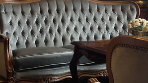 Una habitación con un sofá gris con capitoné, una mesa de centro de madera, un espejo con marco ornamentado, una lámpara y un florero con flores.