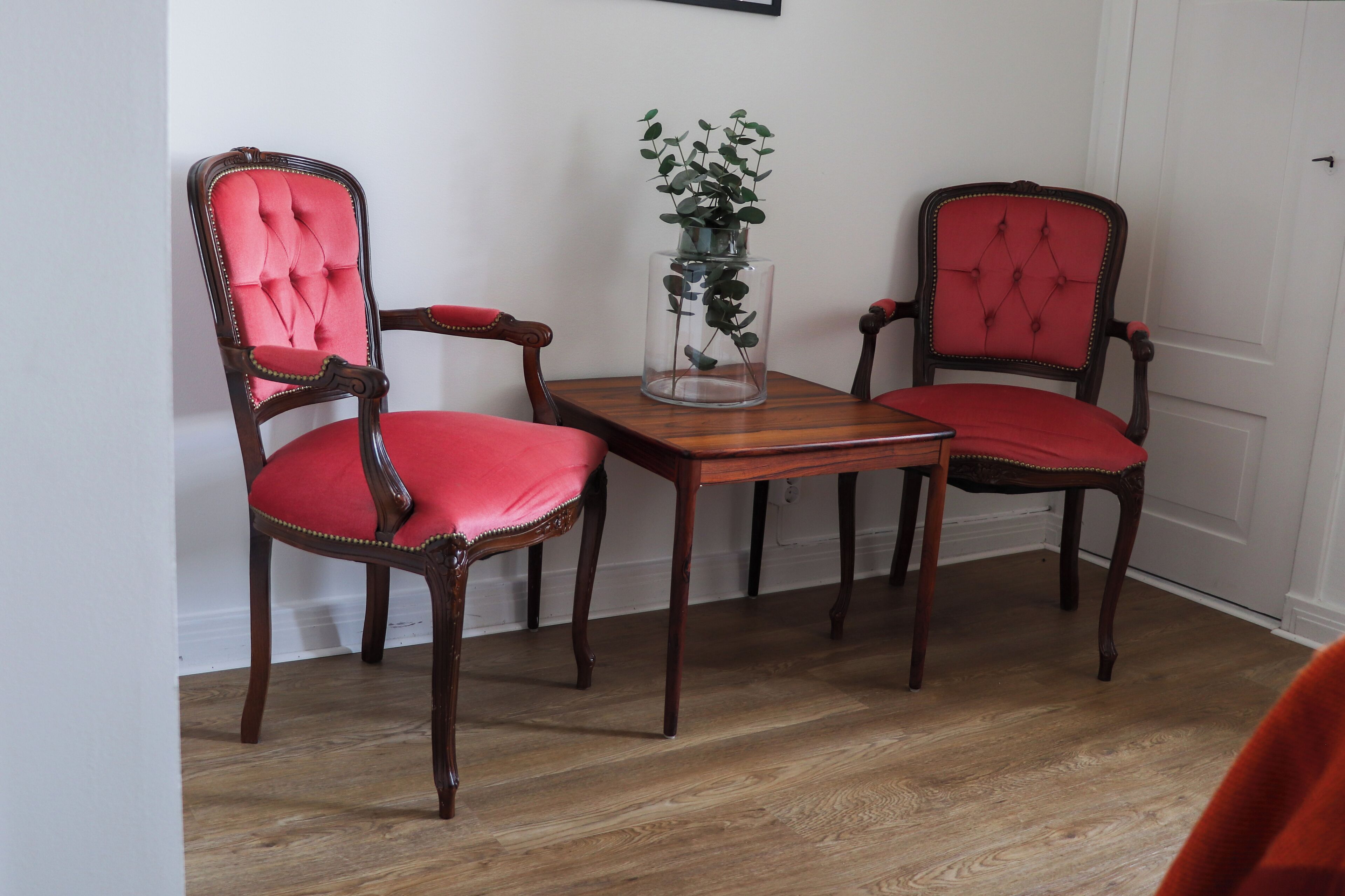 A dining area with two wooden chairs with red upholstery and a glass vase with a plant on a wooden table.