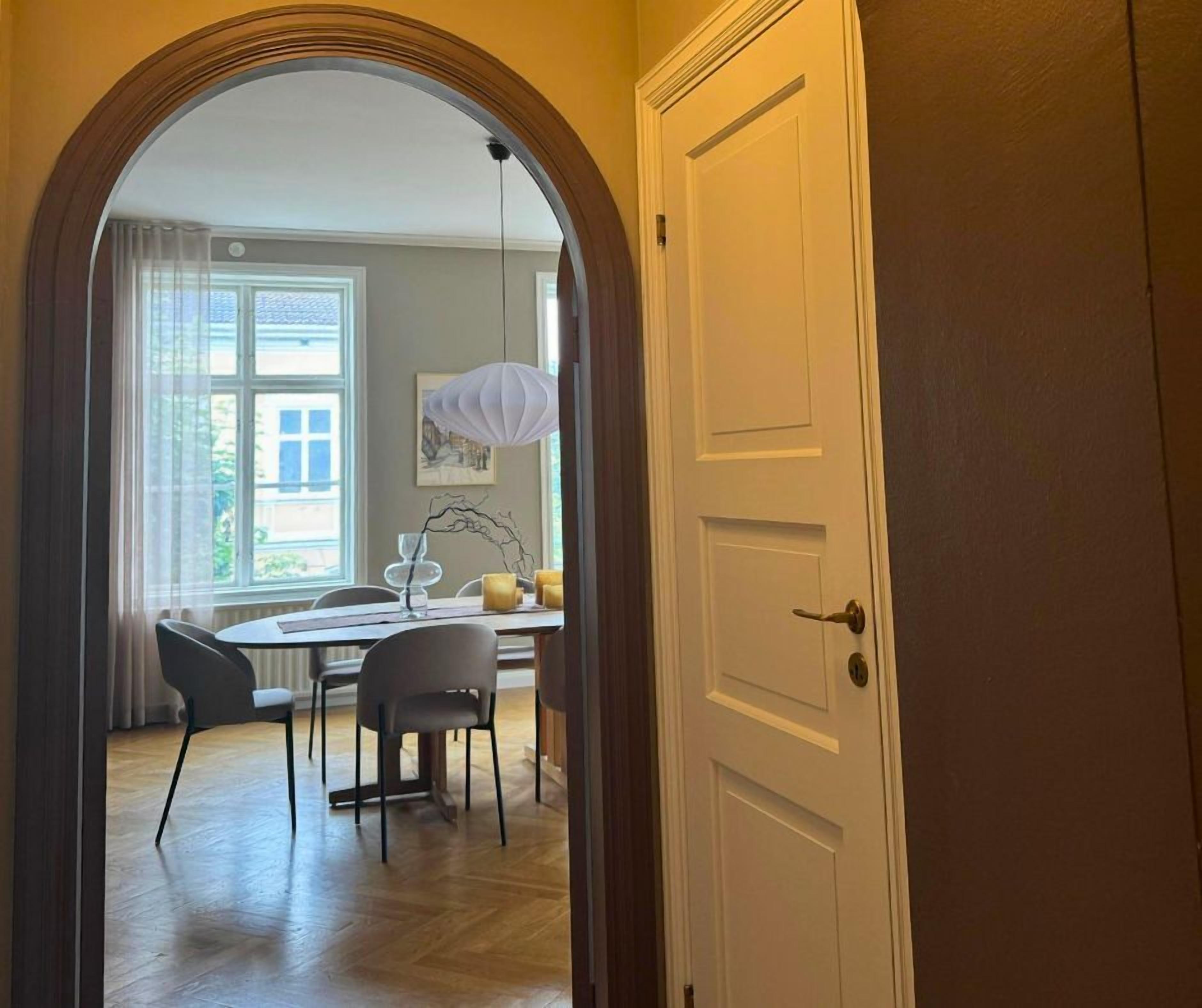 An interior view of a room with a wooden floor, a dining table with chairs, a white door, and a window with curtains.