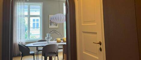 An interior view of a room with a wooden floor, a dining table with chairs, a white door, and a window with curtains.