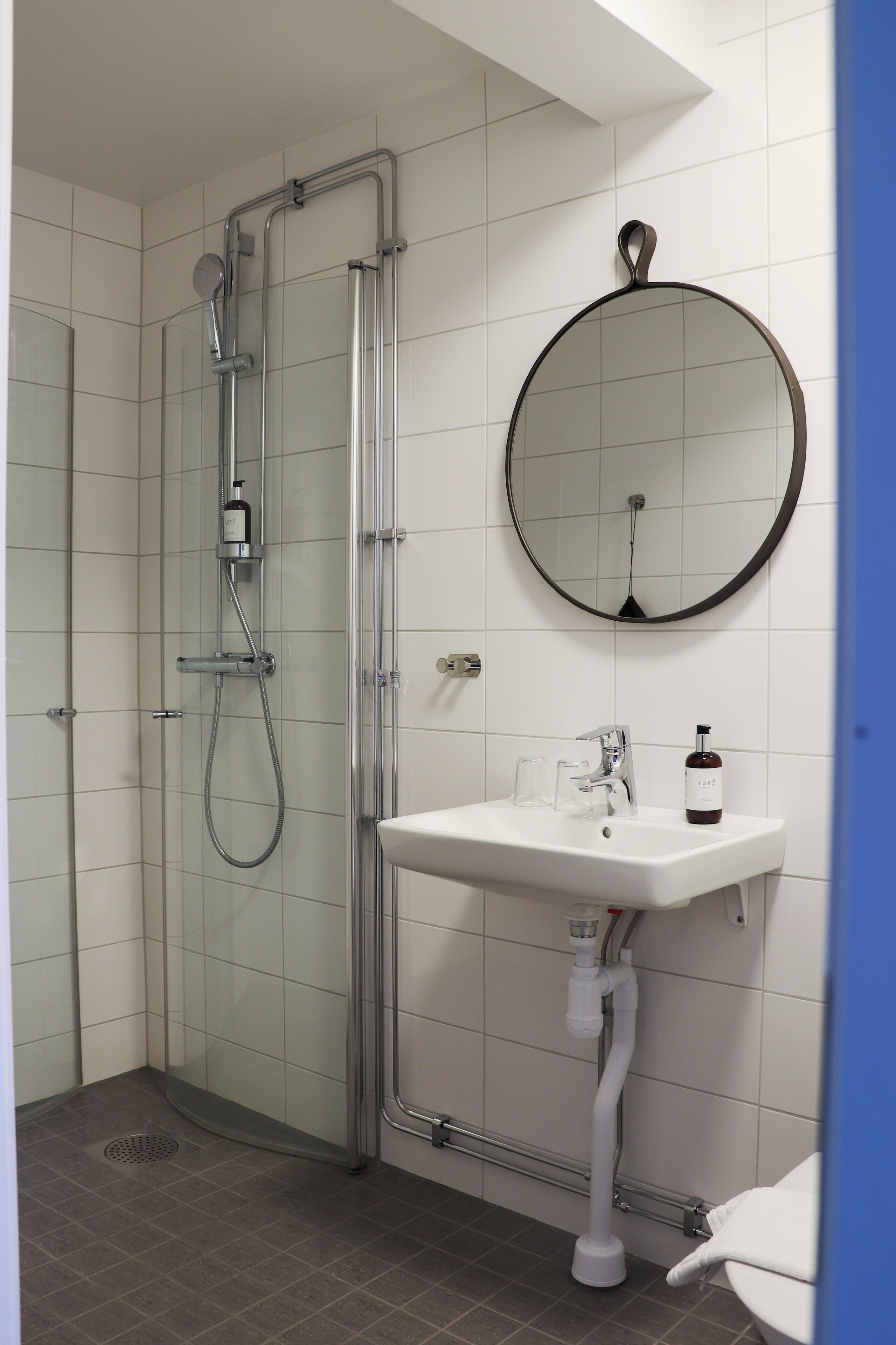 A modern bathroom with a glass-enclosed shower, a round mirror, and a white sink with a bottle of soap.
