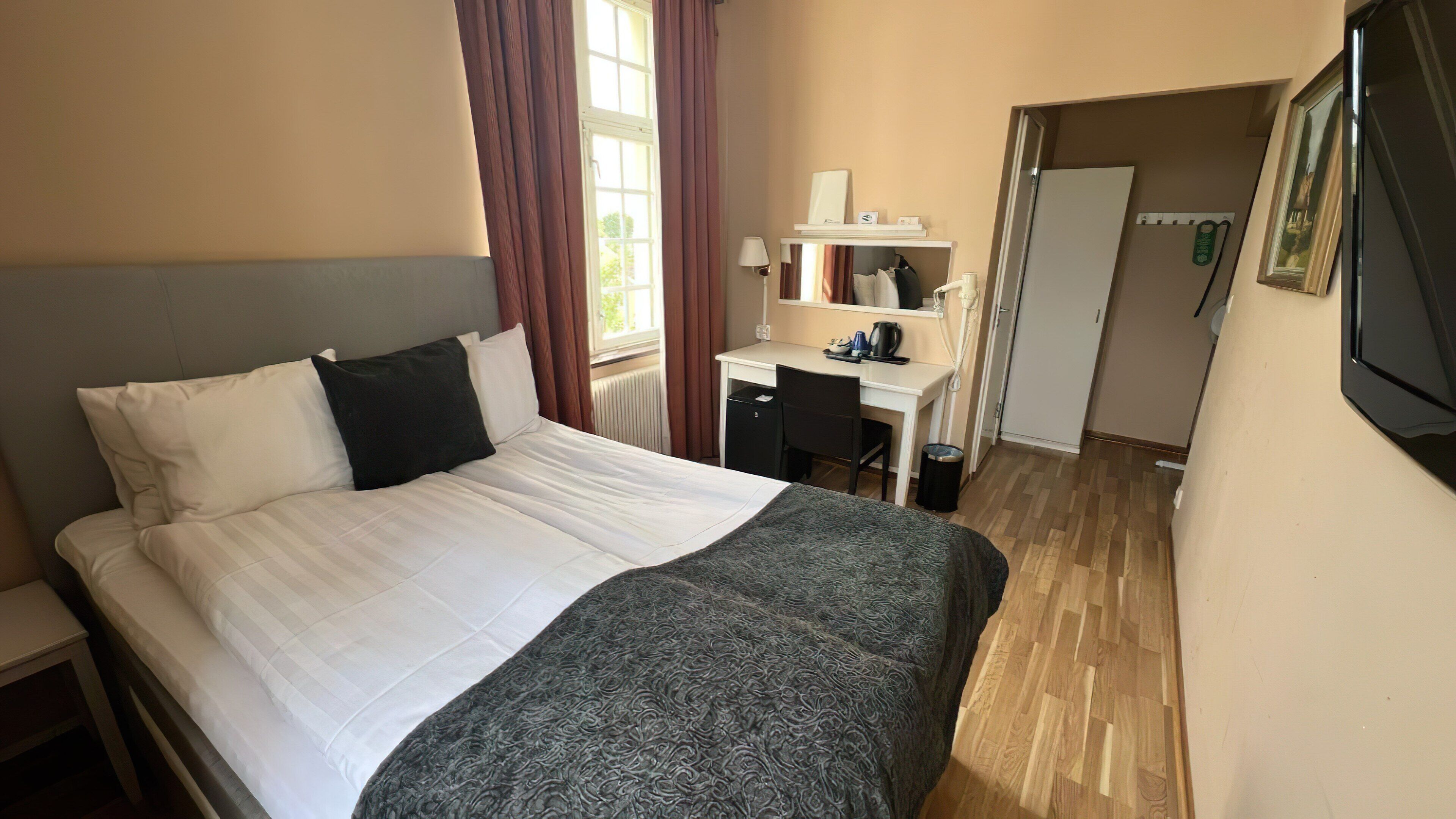 A neatly made bed with white linens and a dark throw blanket, a small desk with a chair, a radiator, and a door leading to another room.