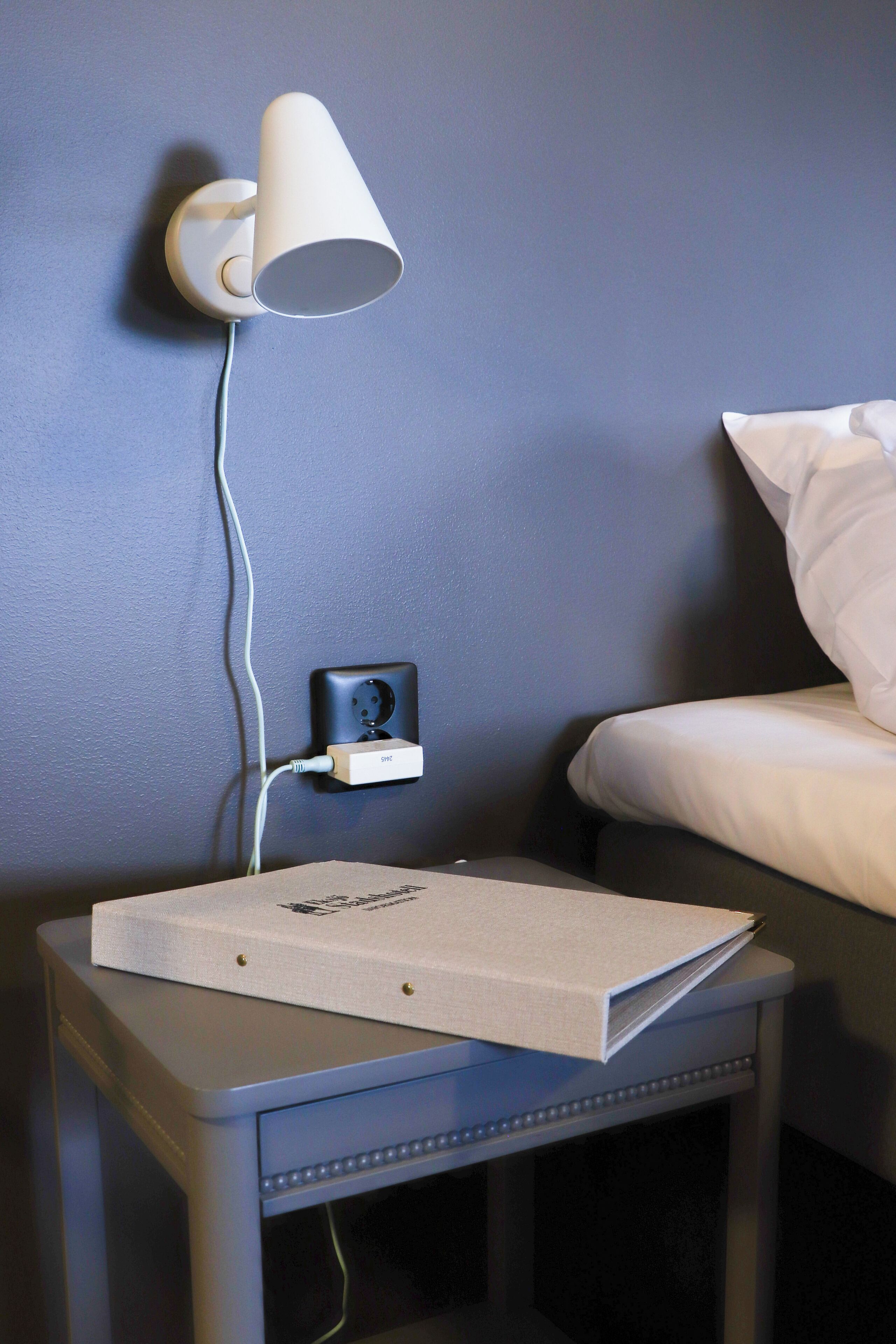 A small bedside table with a lamp, a power outlet, and a box on it.