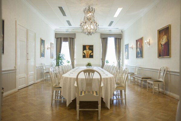 A formal dining room with a round table, white chairs, chandeliers, and framed artwork on the walls. - Eksjo Stadshotell (Eksjo)