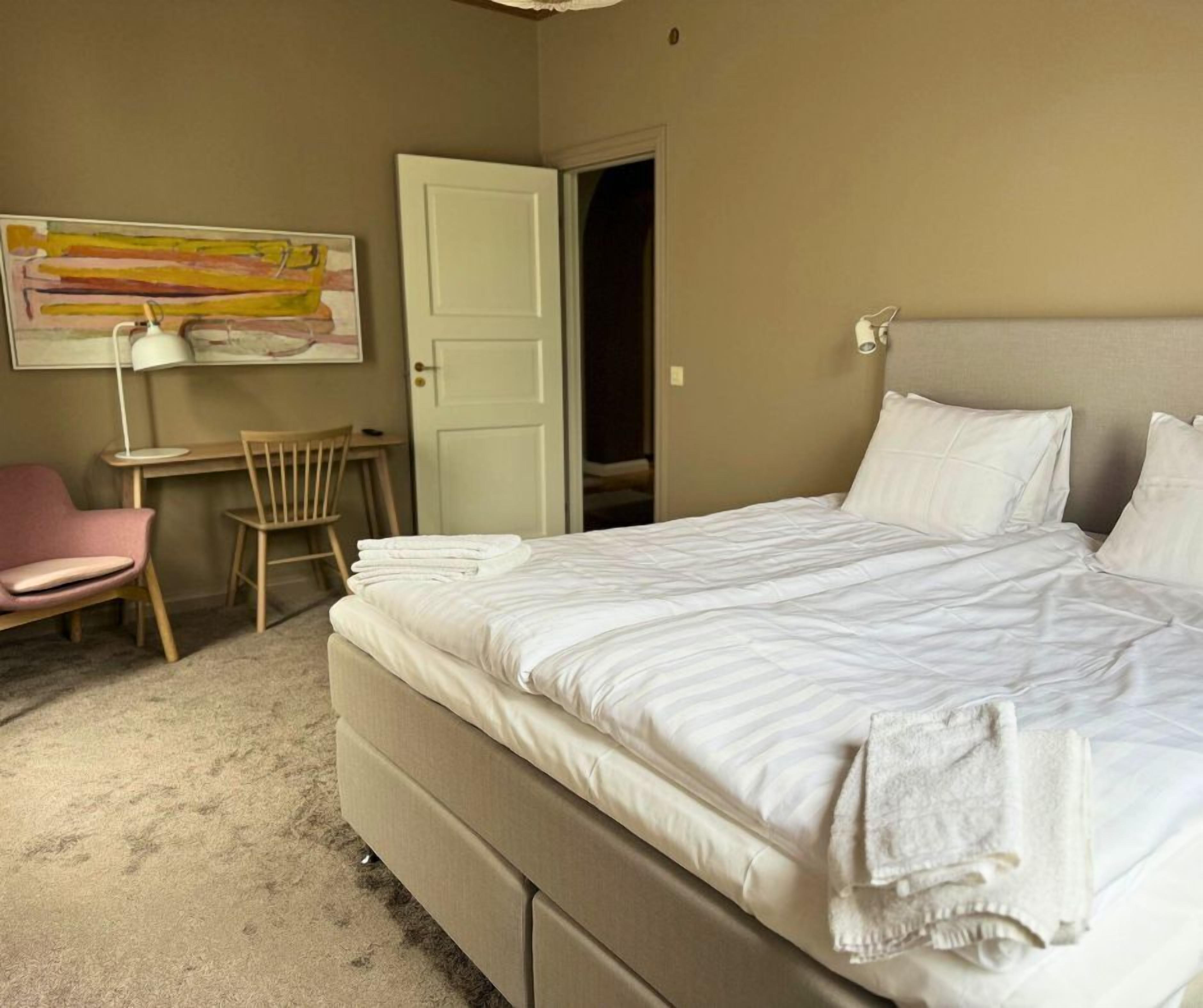 A neatly made bed with white linens, a chair, a desk, and a door leading to another room.