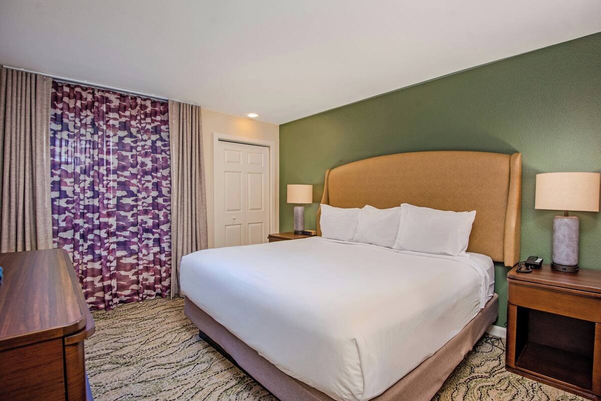 A hotel room with a large bed, two bedside tables with lamps, a wooden nightstand, and a patterned curtain.