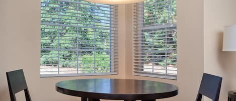 A dining area with a round table and two chairs, large windows with blinds, and a hanging light fixture.