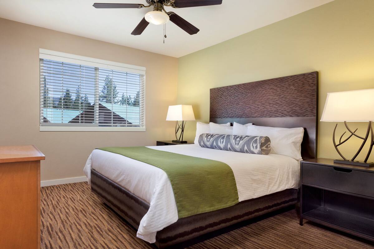 A hotel room with a bed, a nightstand, a lamp, a ceiling fan, and a window with blinds.