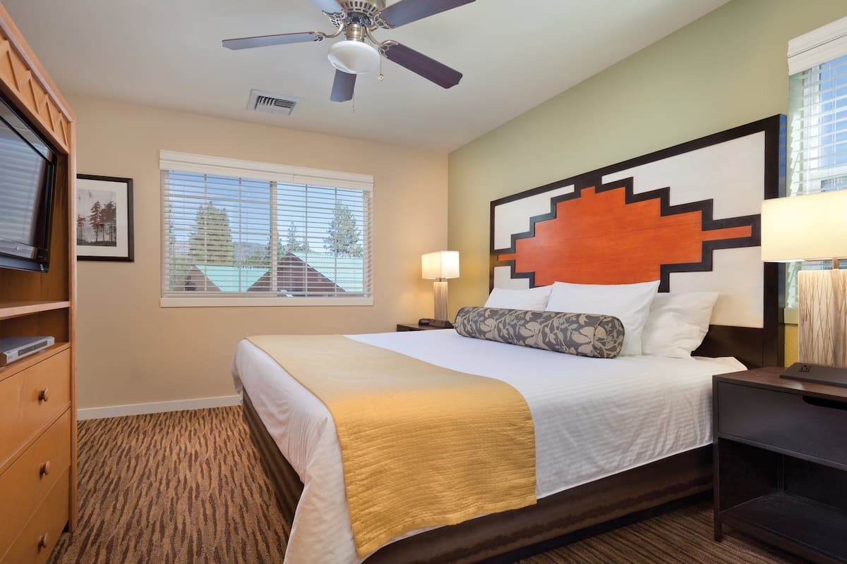 A hotel room with a bed, a ceiling fan, a window with blinds, a nightstand, a lamp, and a dresser.