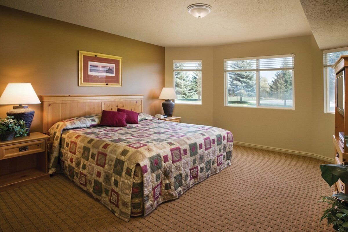 A bedroom with a large bed, two bedside tables with lamps, a headboard, and two windows with blinds.
