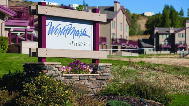 A sign for 'Wynham' by Wyndham, surrounded by a stone planter with flowers, set against a backdrop of residential buildings and a clear sky. - WorldMark Bear Lake (Garden City)