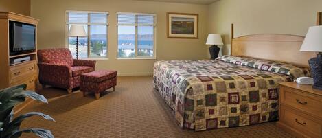 A hotel room with a bed, a chair, a TV, and a view of mountains through the windows.