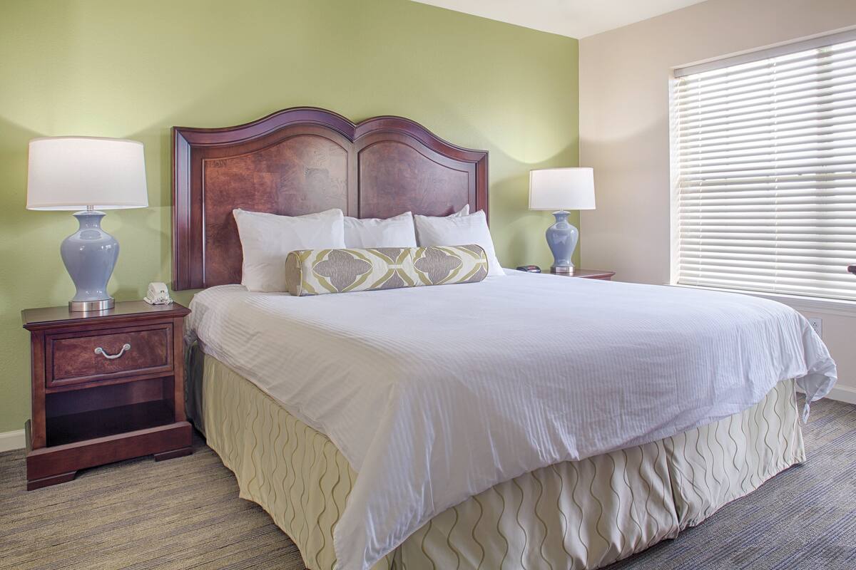 A neatly made bed with a wooden headboard, two bedside tables with lamps, and a window with blinds.