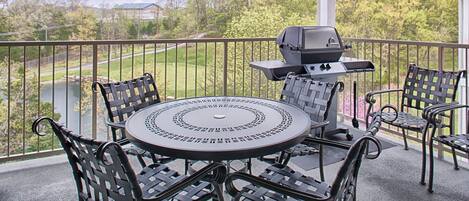 A covered outdoor patio with a round table and four chairs, a grill, and a view of a lake and trees.
