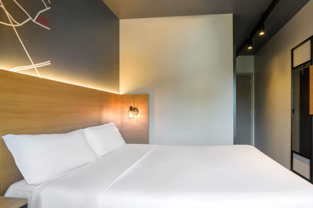 A modern hotel room with a large bed, a wooden headboard, and a wall-mounted light fixture.