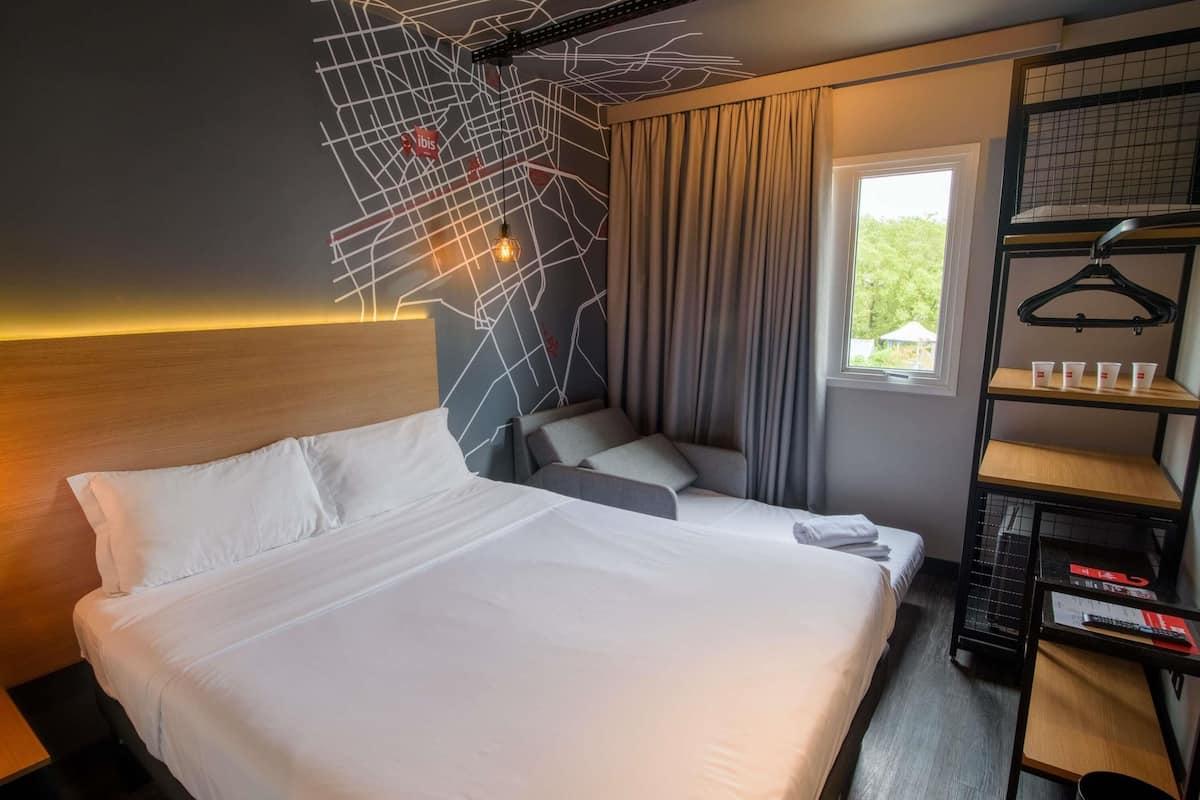 A hotel room with a large bed, a seating area with a sofa and pillows, a window with curtains, a wall-mounted light fixture, and a shelf with cups and a hanger.