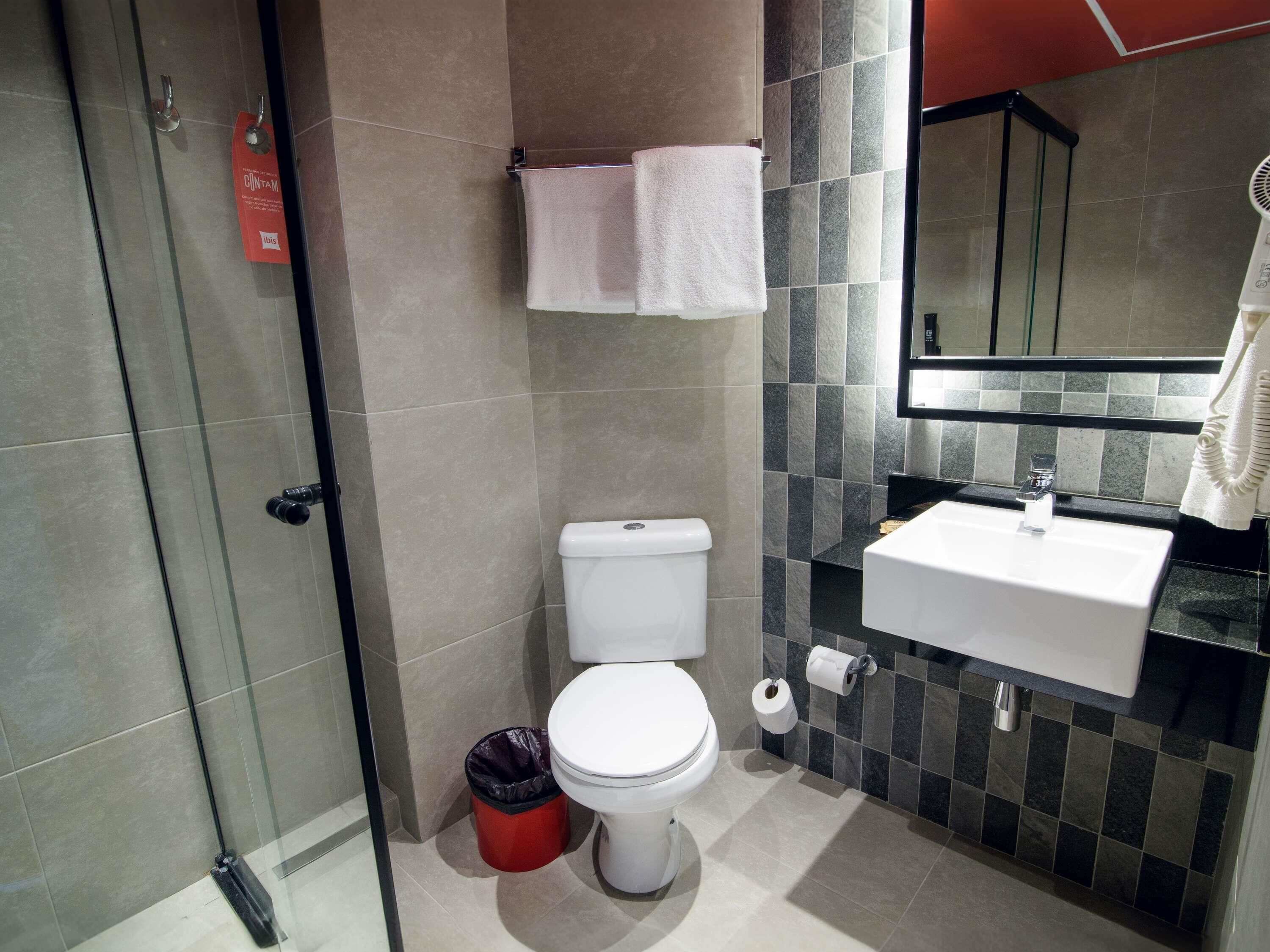 Un baño moderno con cabina de ducha de vidrio, inodoro blanco, cubo de basura negro, lavamanos blanco con espejo y toalla colgada en un perchero.