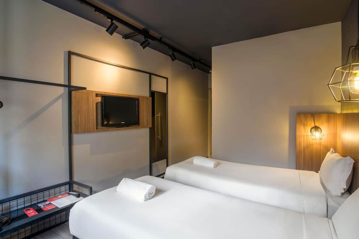 A modern hotel room with a large bed, a flat-screen TV mounted on the wall, a bedside table with a red sign, and a unique geometric light fixture.