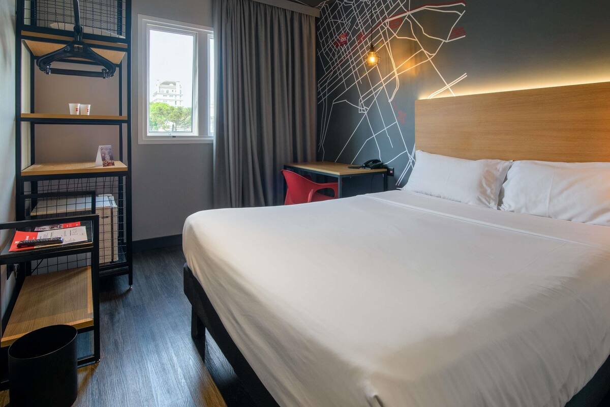 A hotel room with a large bed, a desk, a chair, a shelf with items, and a window with curtains.