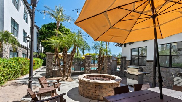 An outdoor patio with a brick fire pit, wooden tables and chairs, a yellow umbrella, and a pool in the background. - Sonesta ES Suites Phoenix Goodyear (Goodyear)