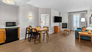A modern living room with a flat-screen TV, a sofa, a dining table with chairs, a kitchen area with a microwave, and a fireplace. - Sonesta ES Suites Phoenix Goodyear (Goodyear)