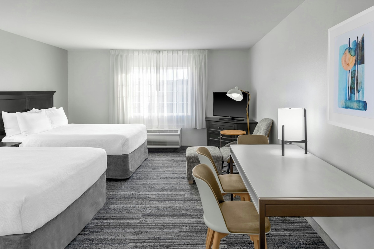 Een hotelkamer met twee bedden, een bureau, een stoel, een televisie en een schilderij aan de muur.