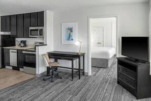 Een kleine hotelkamer met een kitchenette, een bed, een bureau met stoel, een televisie en een schilderij aan de muur.