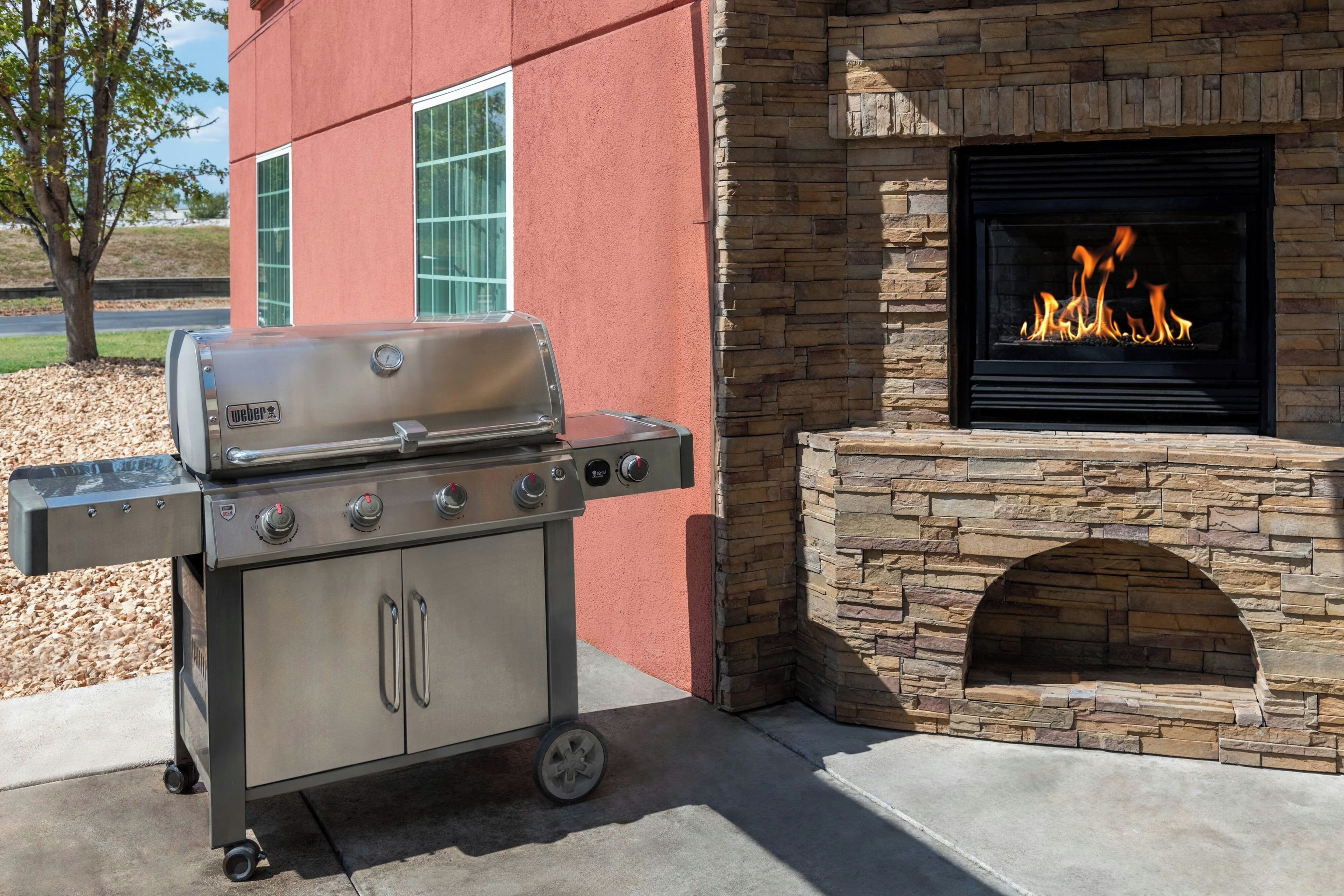 A Weber grill with four burners and a side cabinet, positioned next to a stone fireplace with a visible fire.