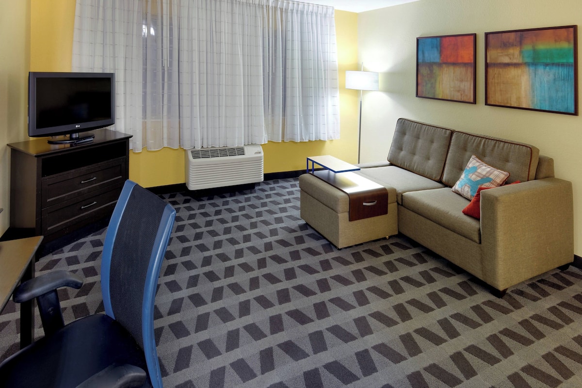 A hotel room with a TV, a sofa, a chair, a small table, and a lamp.