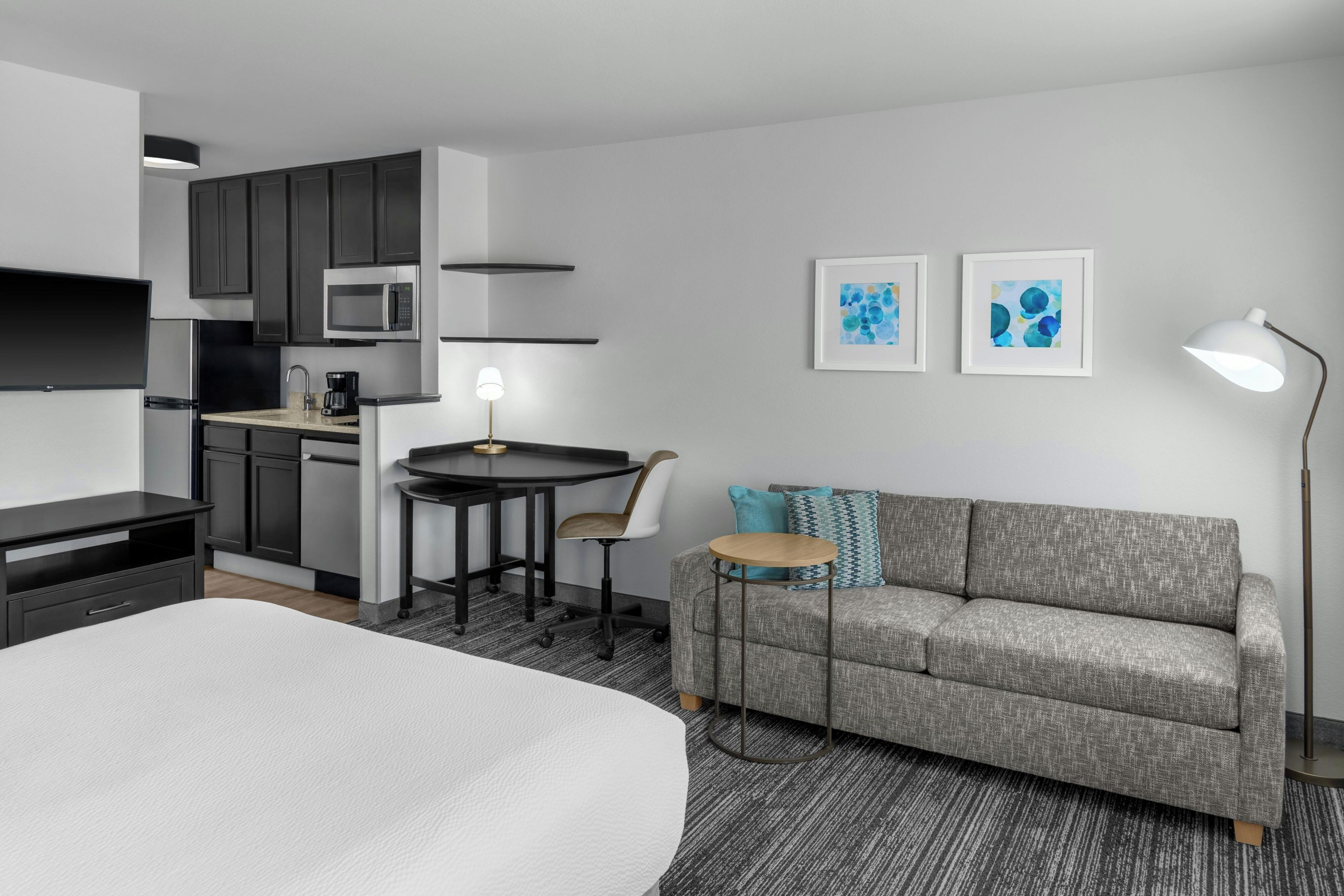 A modern hotel room with a bed, kitchenette, dining area, and a sofa with a small table.