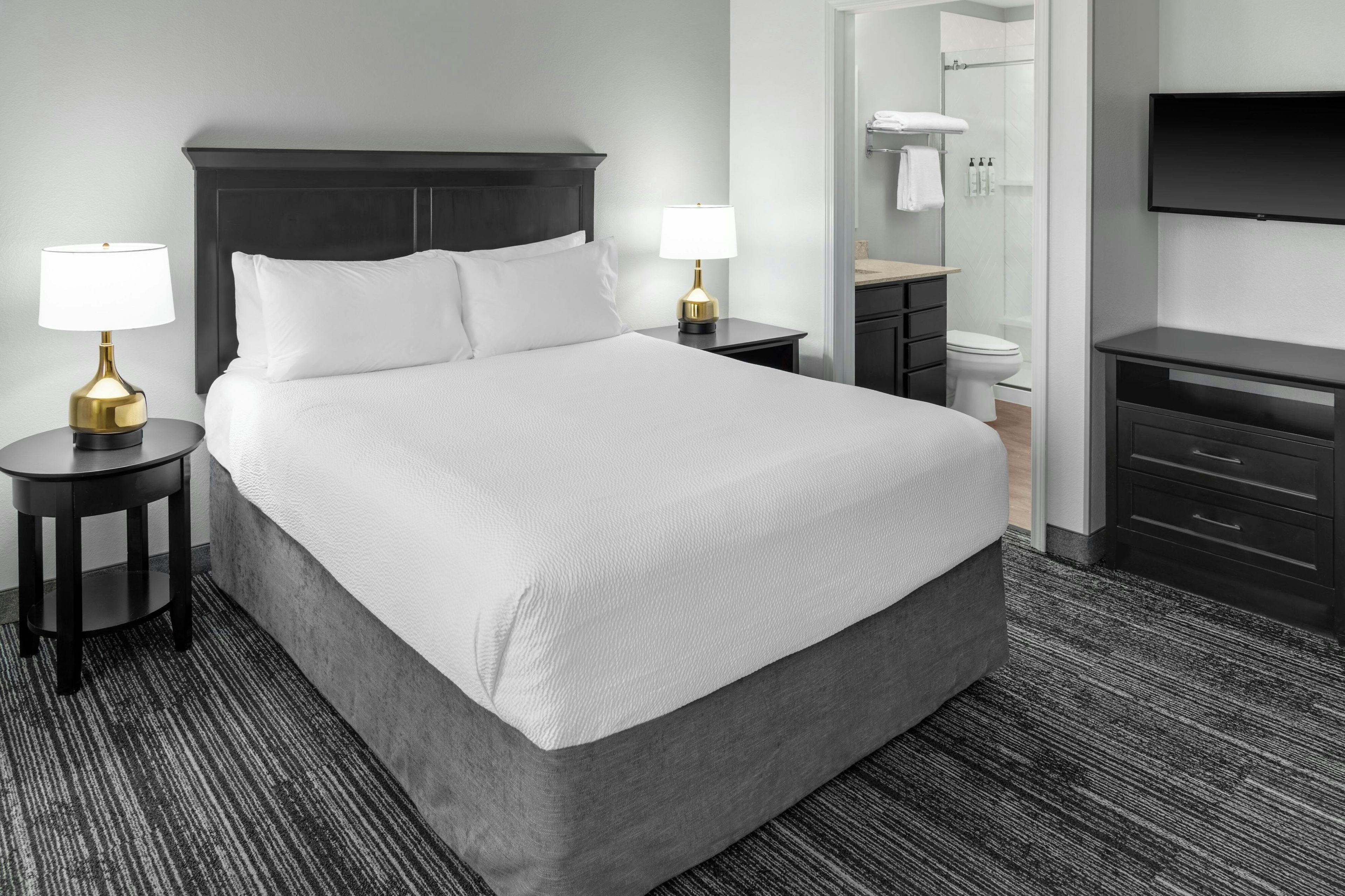 Ein Hotelzimmer mit einem großen Bett, zwei Nachttischen mit Lampen, einem dunklen Holz-Kopfteil, einem Badezimmer inklusive Toilette und Dusche sowie einem an der Wand befestigten Flachbildfernseher.