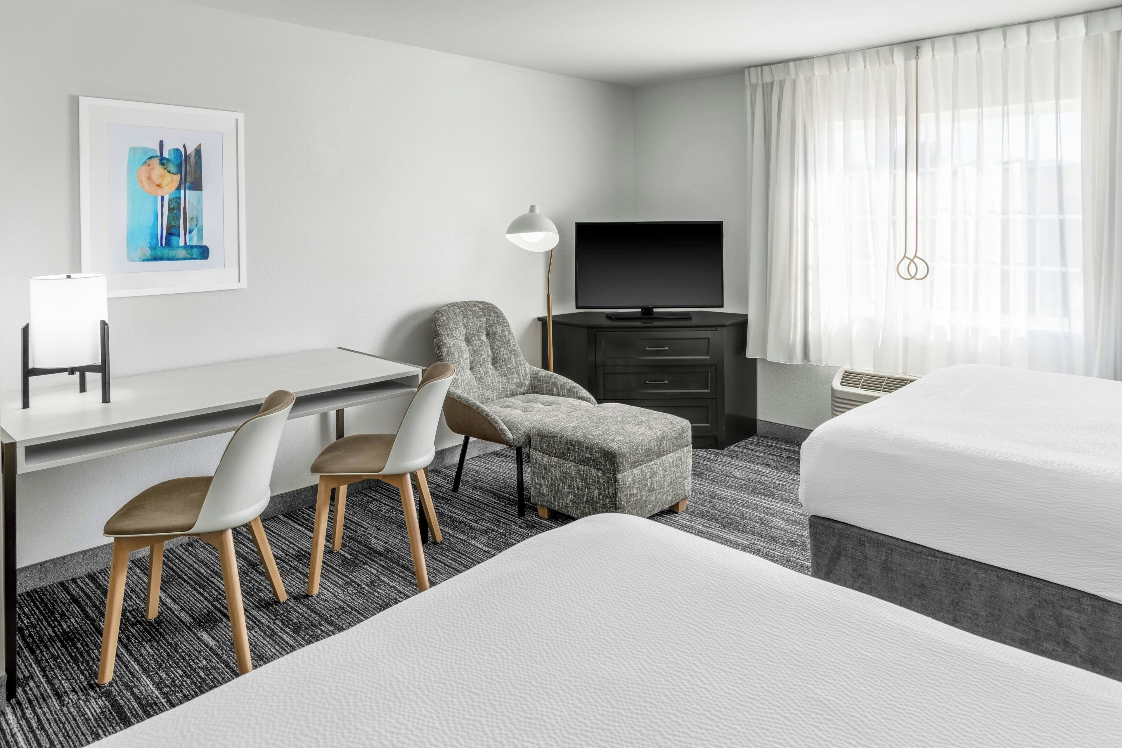 A hotel room with a bed, a desk, a chair, a television, and a painting on the wall.