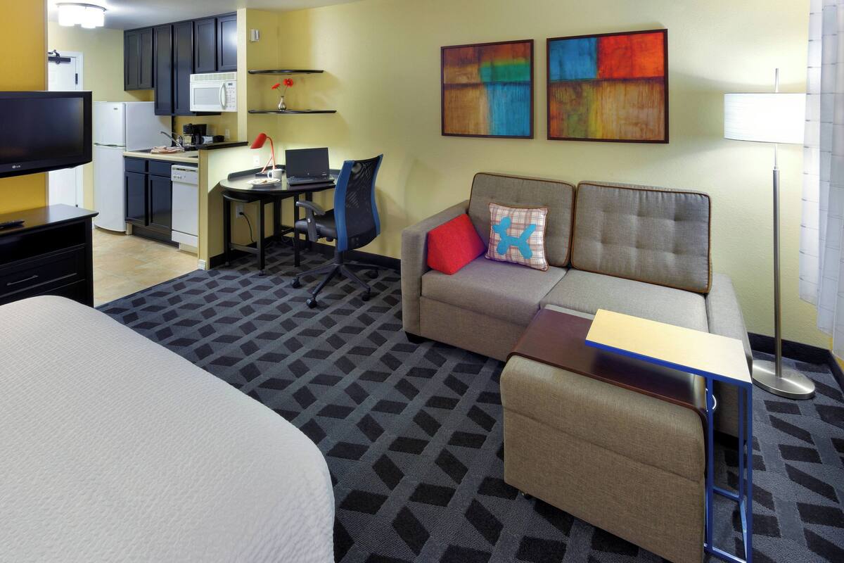 A hotel room with a bed, a sofa, a desk with a laptop, a microwave, a TV, and artwork on the wall.