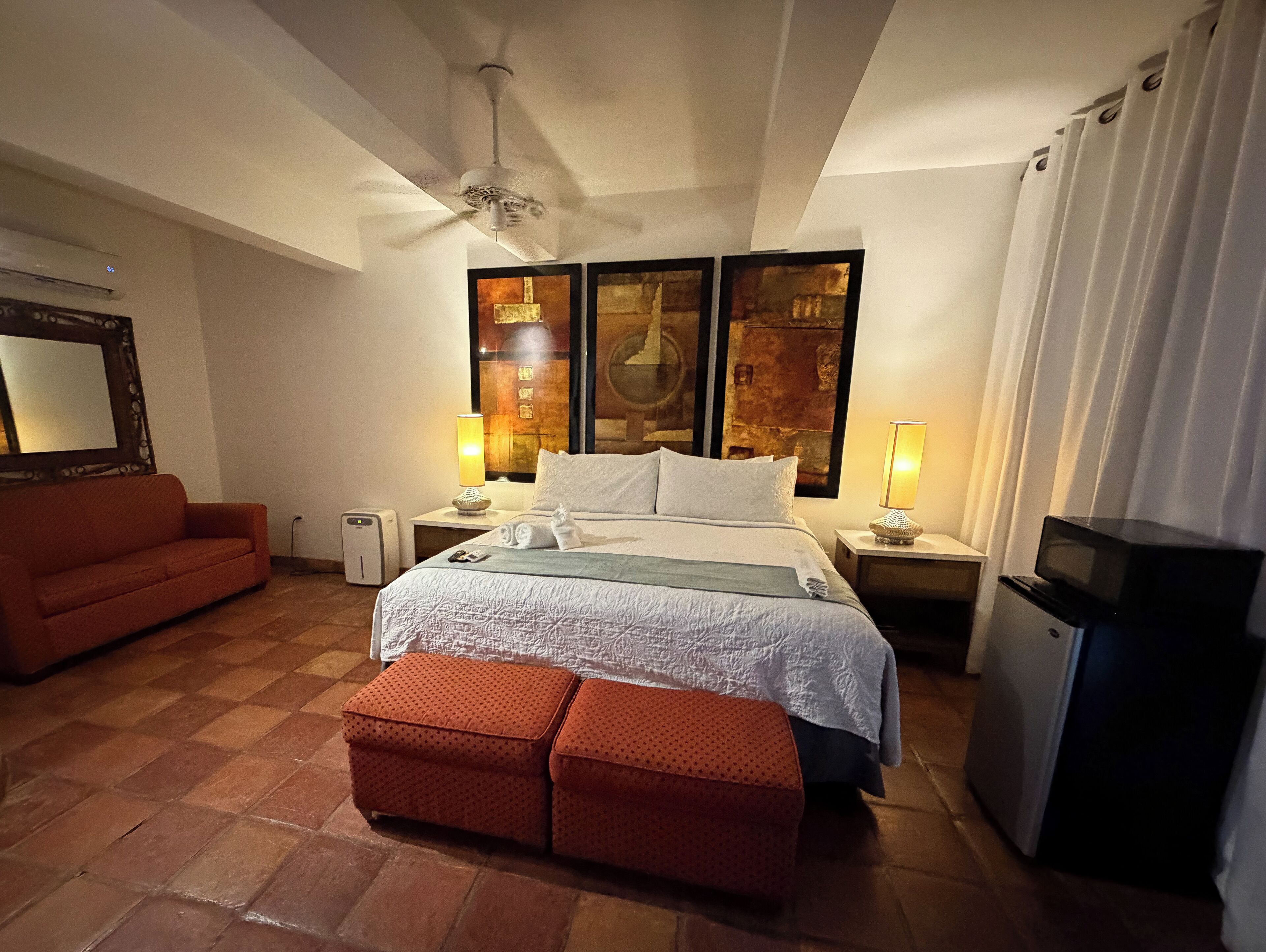 Una habitación de hotel con cama, sofá rojo, una mesita, televisor y ventilador de techo.