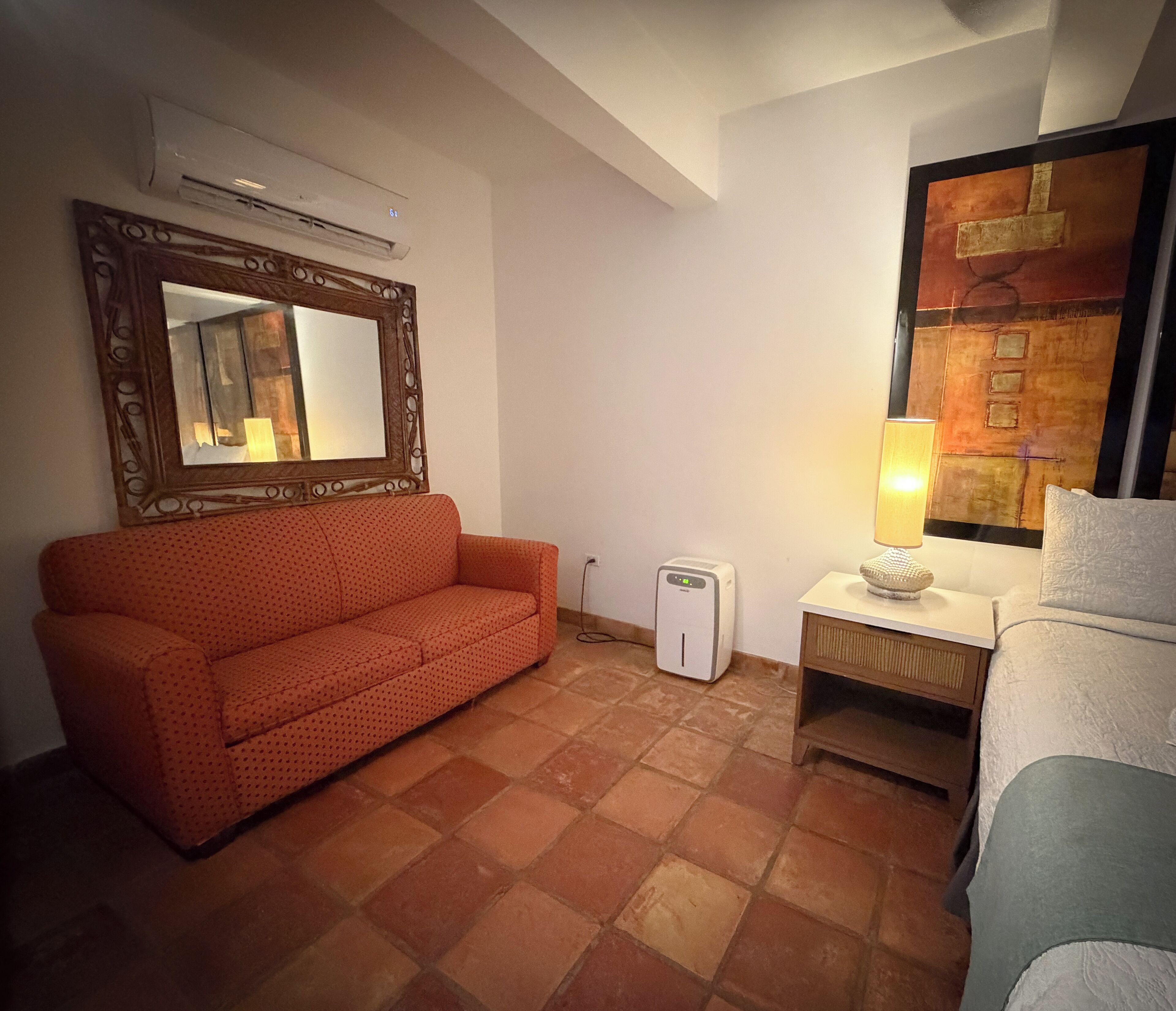 Una habitación con un sofá rojo, una mesita de noche blanca con lámpara, un pequeño calefactor blanco y un cuadro abstracto grande en la pared.