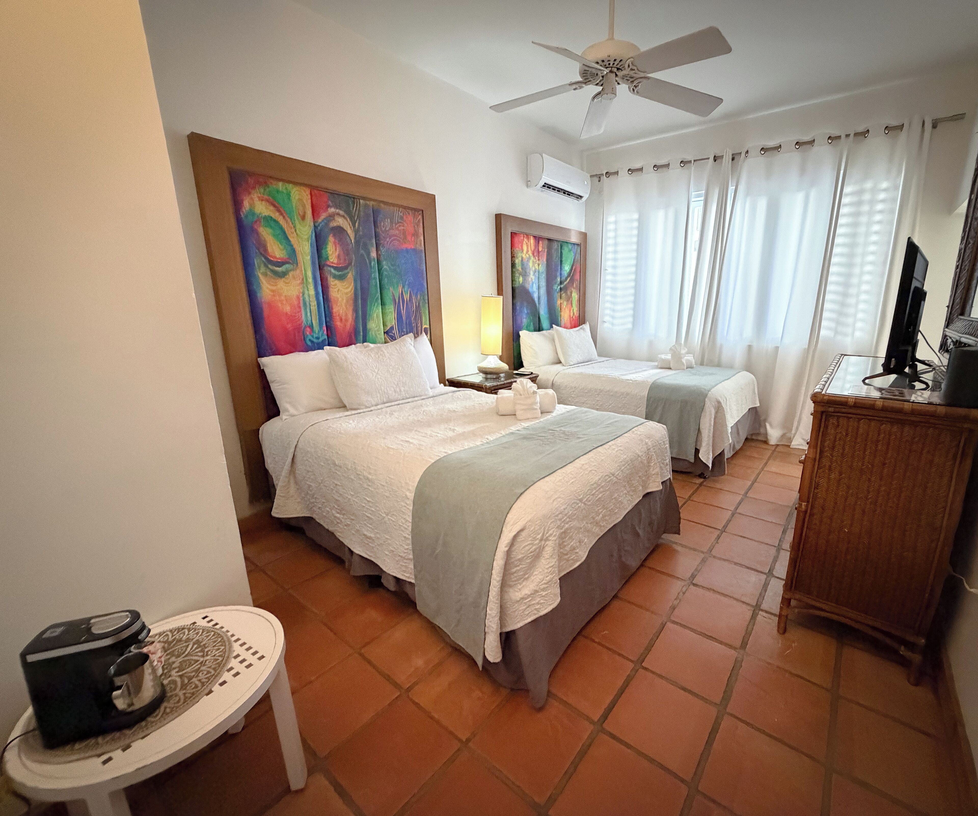 Habitación de hotel con dos camas, ventilador de techo, aire acondicionado, televisor y una mesita con cafetera.