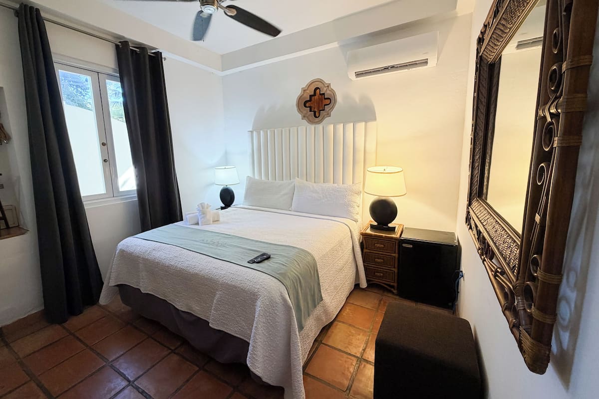 Une chambre d’hôtel avec un lit, un ventilateur de plafond, une table de chevet avec une lampe, un miroir et une fenêtre avec des rideaux.