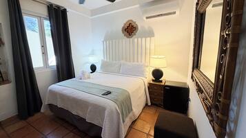 Una habitación de hotel con cama, ventilador de techo, mesita de noche con lámpara, espejo y ventana con cortinas.