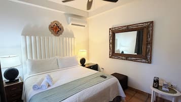 Una cama bien hecha con cabecera blanca, un ventilador de techo, una mesita de noche con lámpara, una pequeña mesa con cafetera y un espejo en la pared.