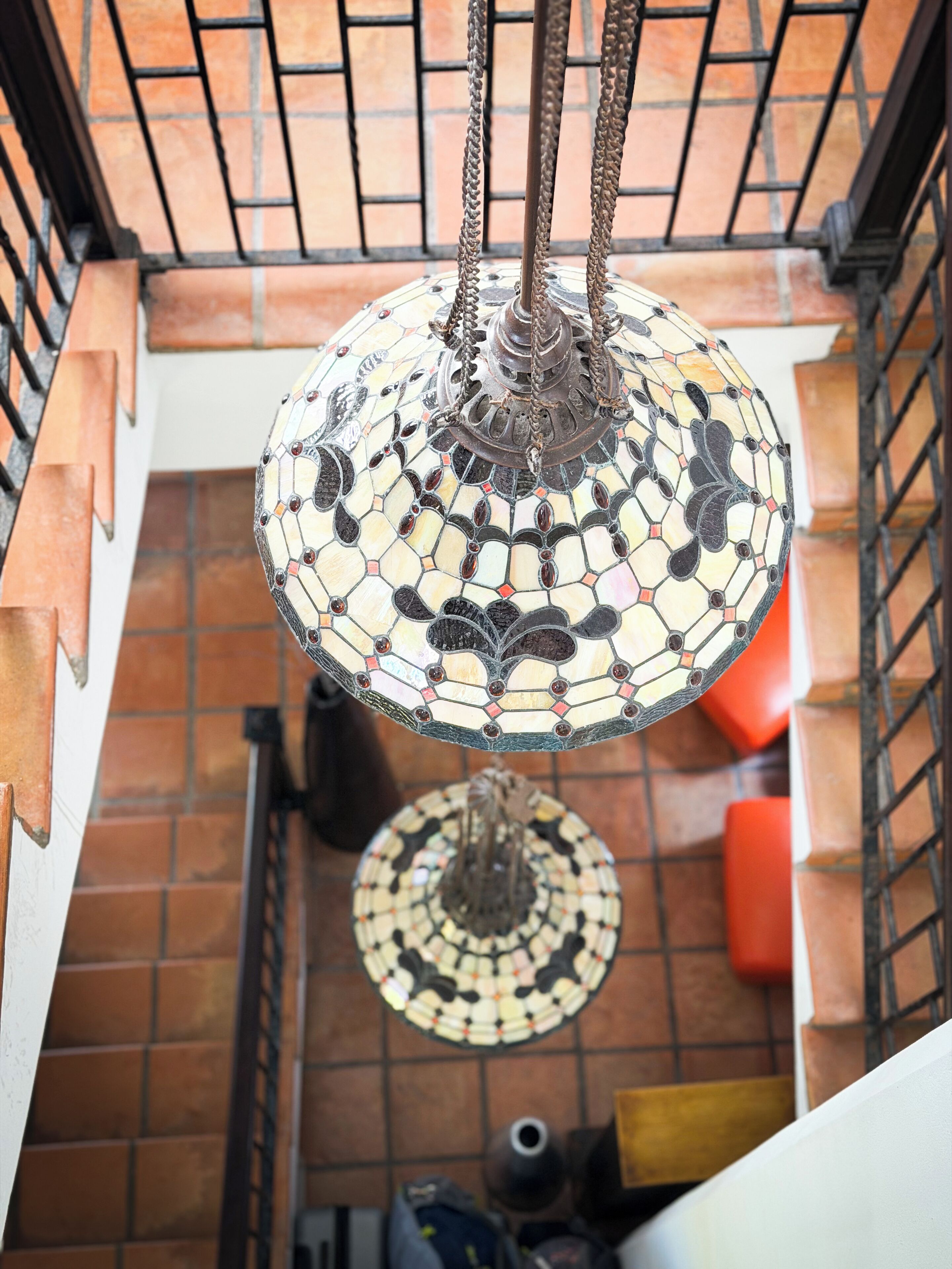 Una lámpara colgante de mosaico con base de metal y cadenas, suspendida del techo azulejado en una habitación con suelo de azulejos y una escalera.