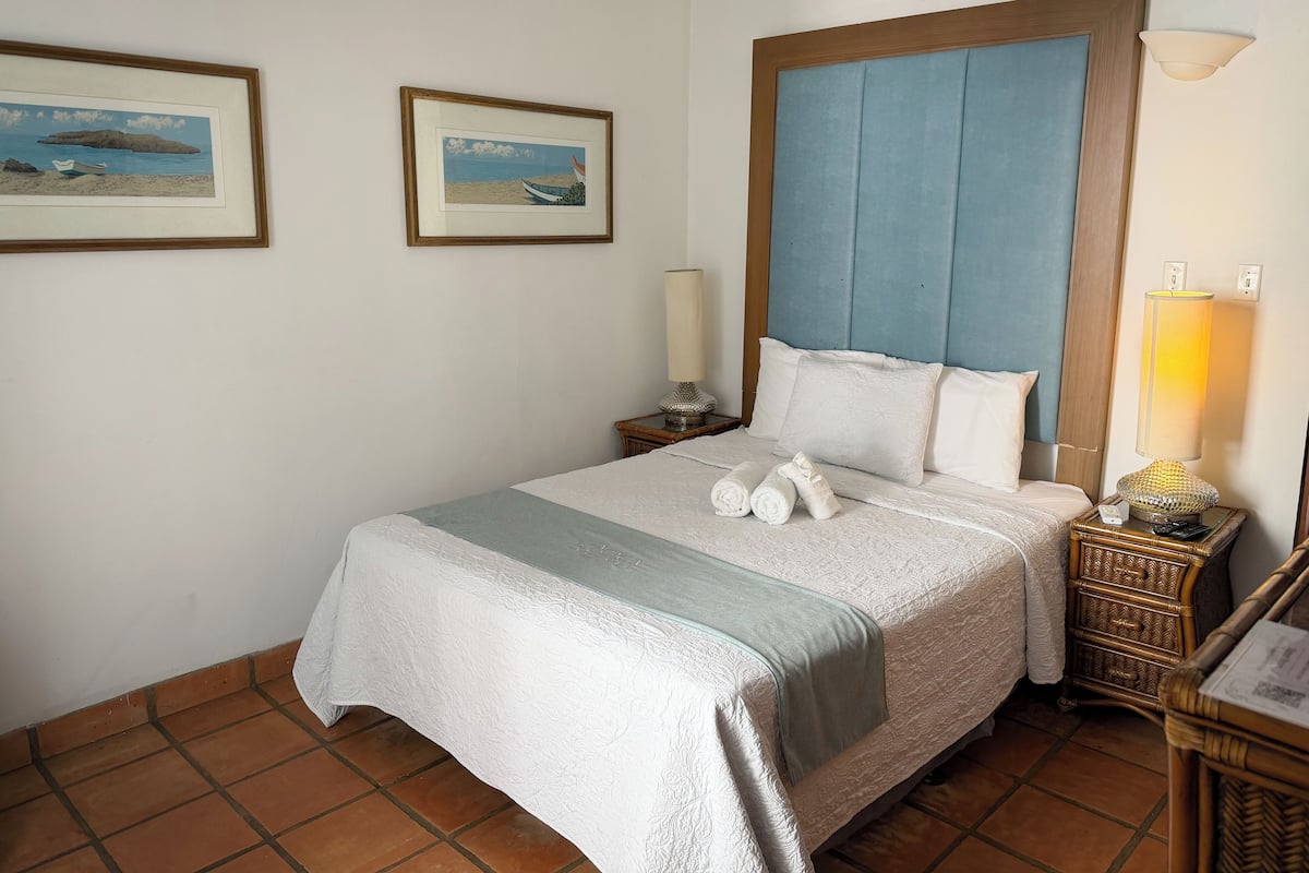 Un lit bien fait, avec une tête de lit bleue, deux tableaux encadrés accrochés au mur et une lampe de chevet.