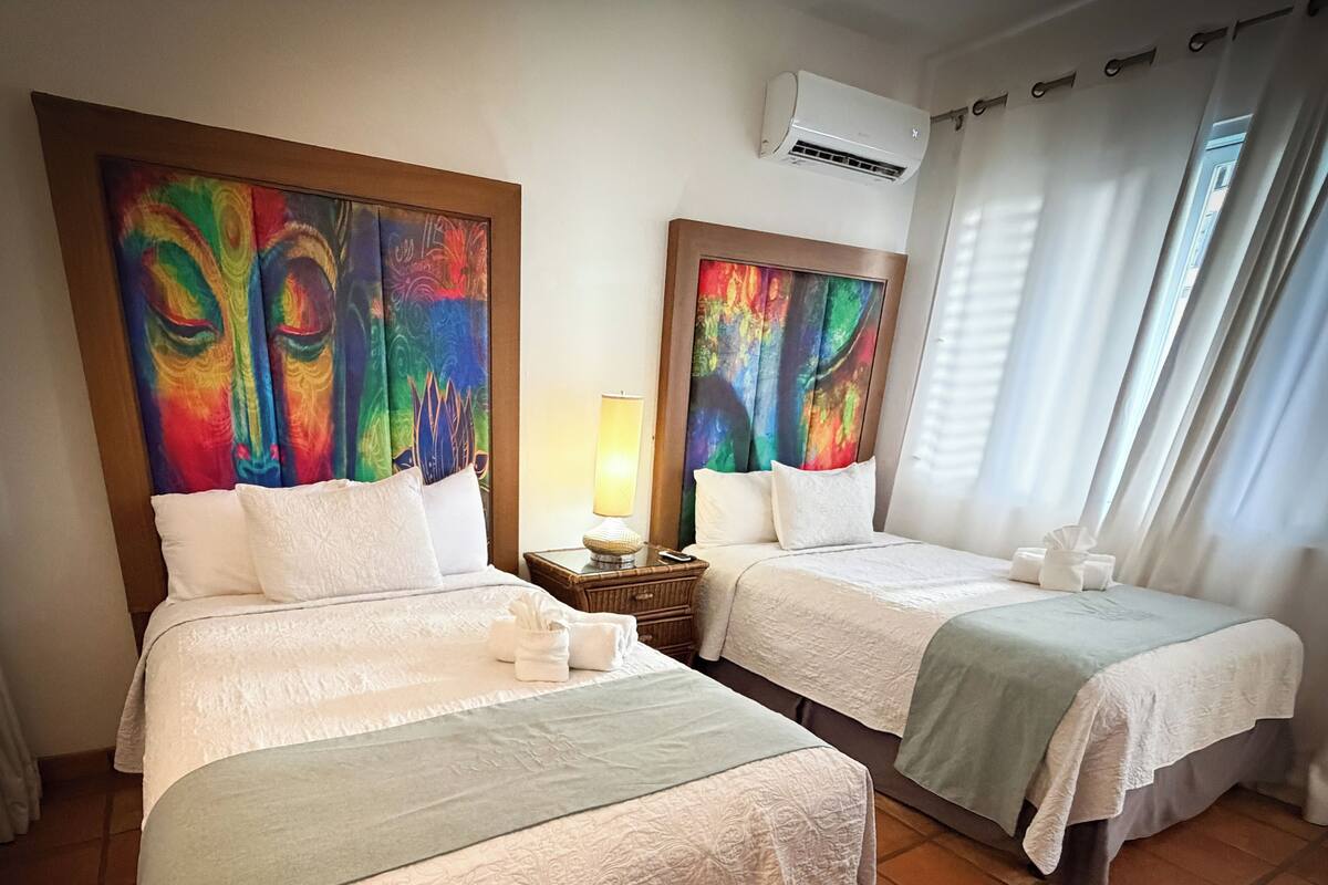 Une chambre d’hôtel avec deux lits, chacun surmonté d’un tableau abstrait aux couleurs vives, une lampe de chevet et une fenêtre avec des rideaux.