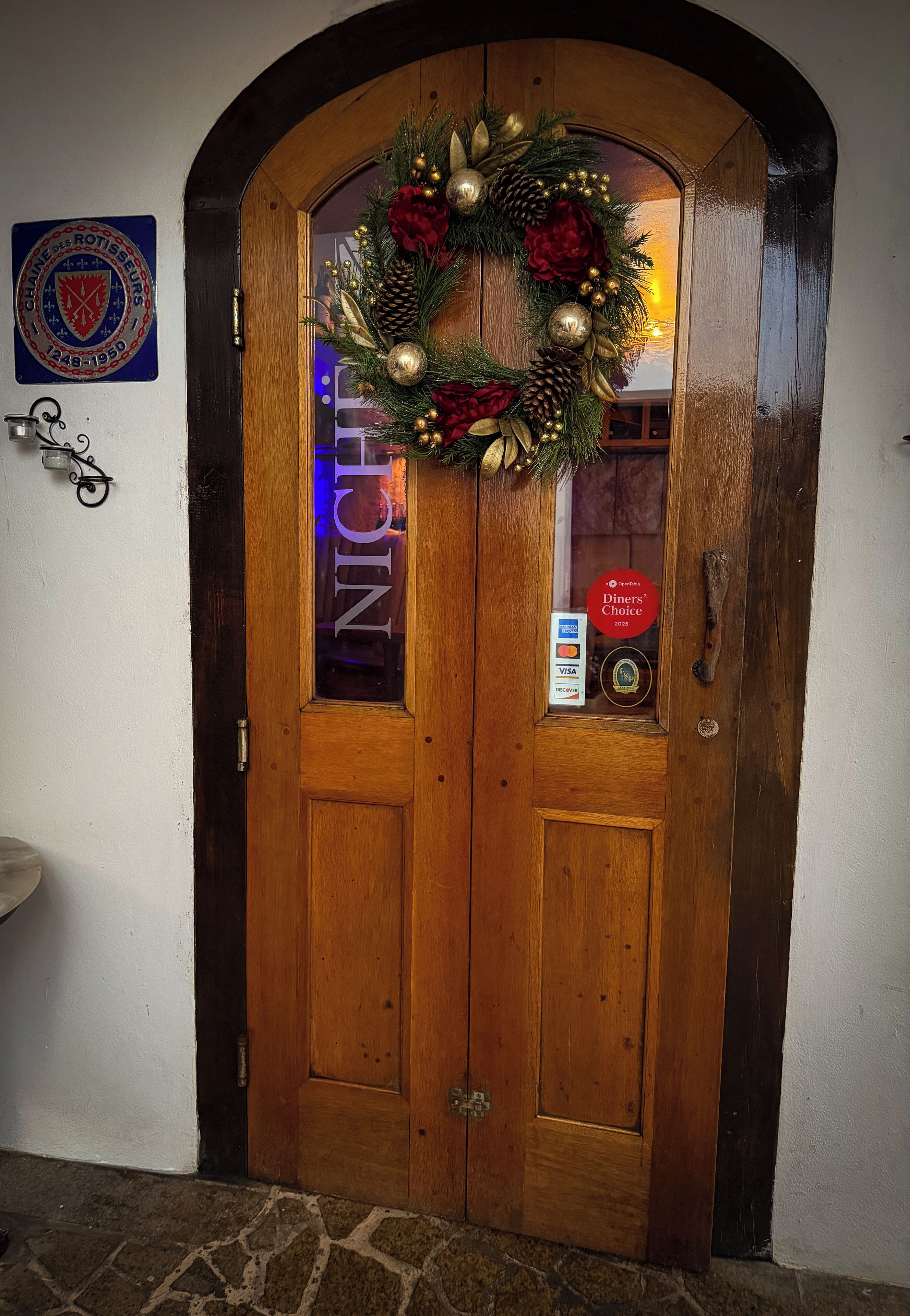 Une porte à double battant en bois, ornée d’une guirlande festive, d’une enseigne « Diners Choice » et d’une plaque « Château de Rotisseurs ».