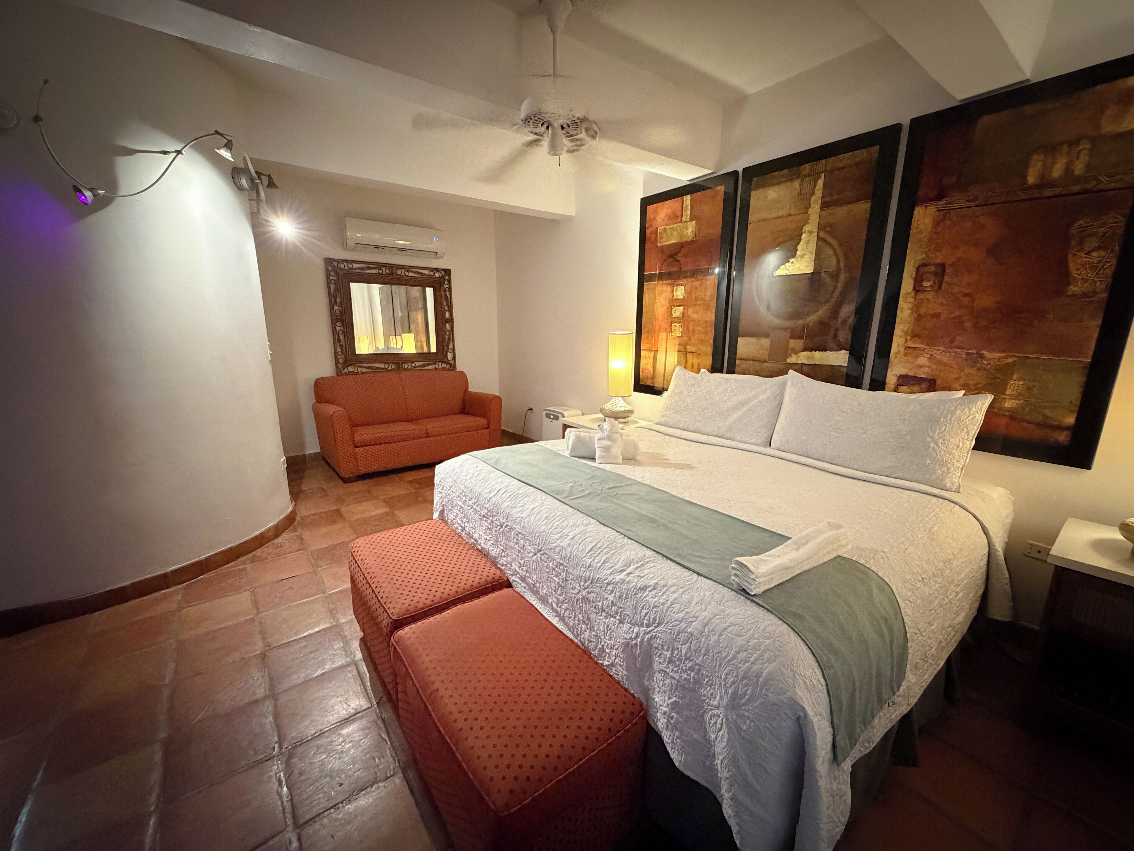 A bedroom with a bed, a sofa, a ceiling fan, an air conditioning unit, and a wall with artwork.