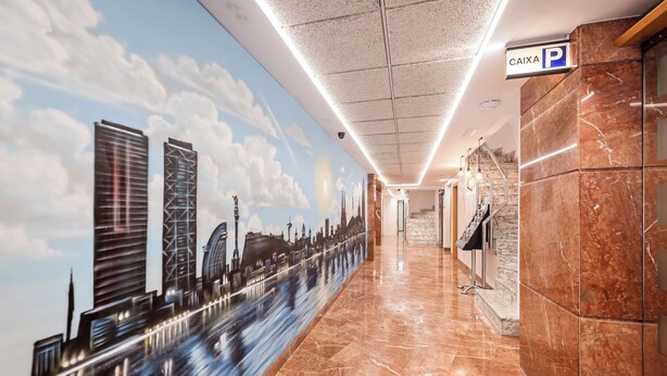 Pasillo de hotel con un mural de un paisaje urbano, suelo de mármol y un cartel que indica una zona de estacionamiento.