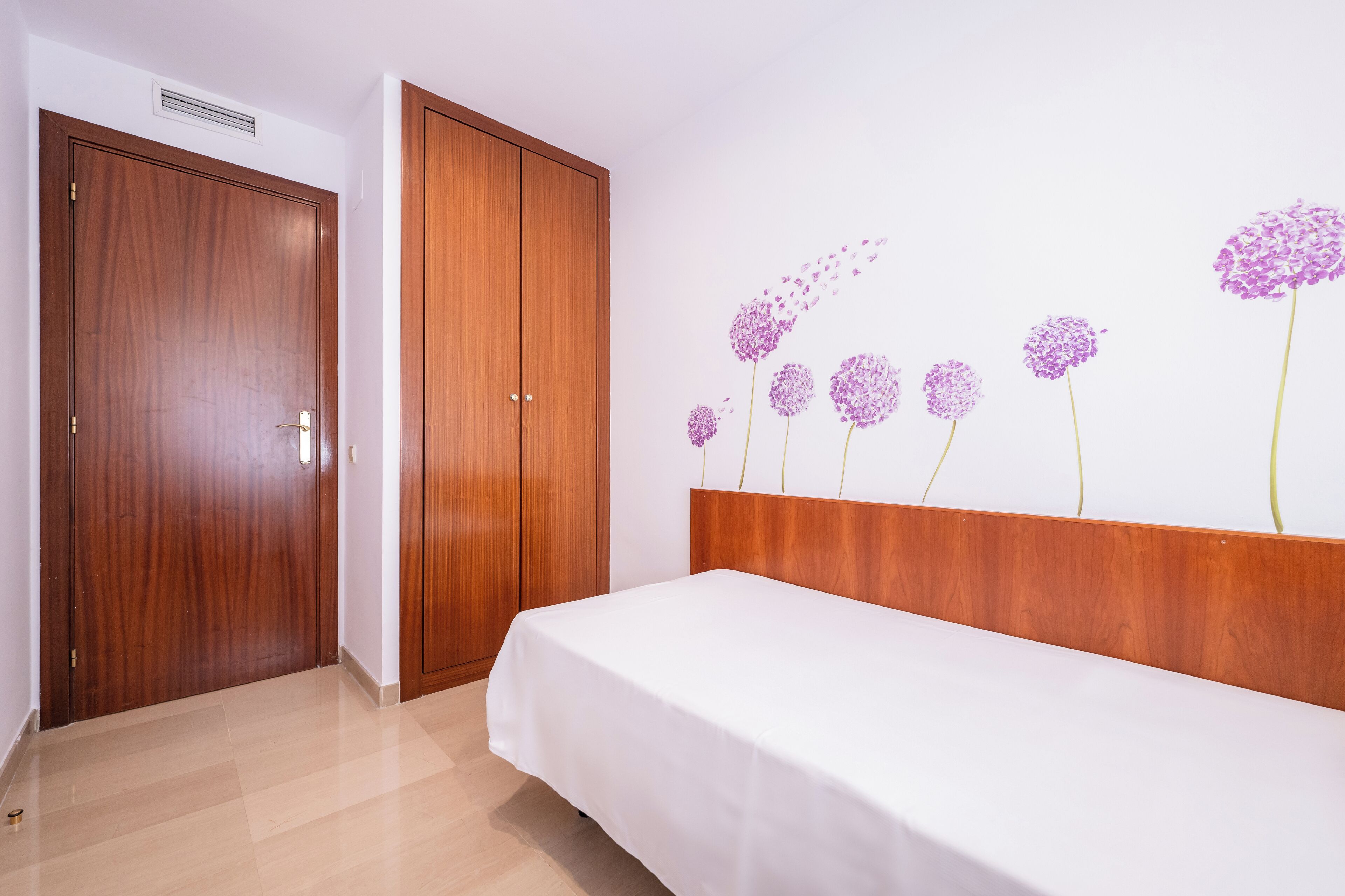 Ein Schlafzimmer mit einem Holzschrank, einem Bett mit weißer Bettdecke und einer Wand mit floralem Wandbild.