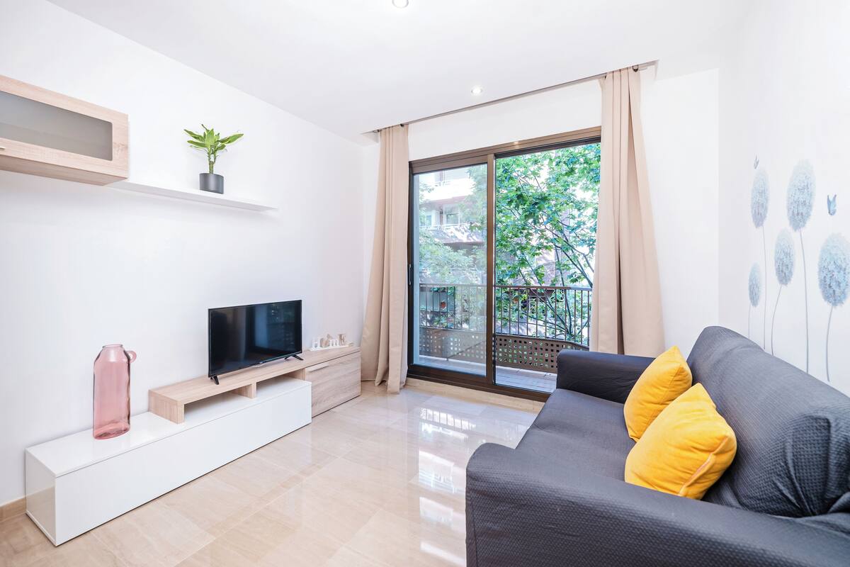A modern living room with a flat-screen TV, a grey sofa with yellow cushions, a white console table, a potted plant, and a sliding glass door leading to a balcony.