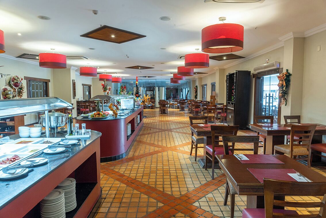 Espaço de refeições de hotel com buffet, várias mesas e cadeiras.