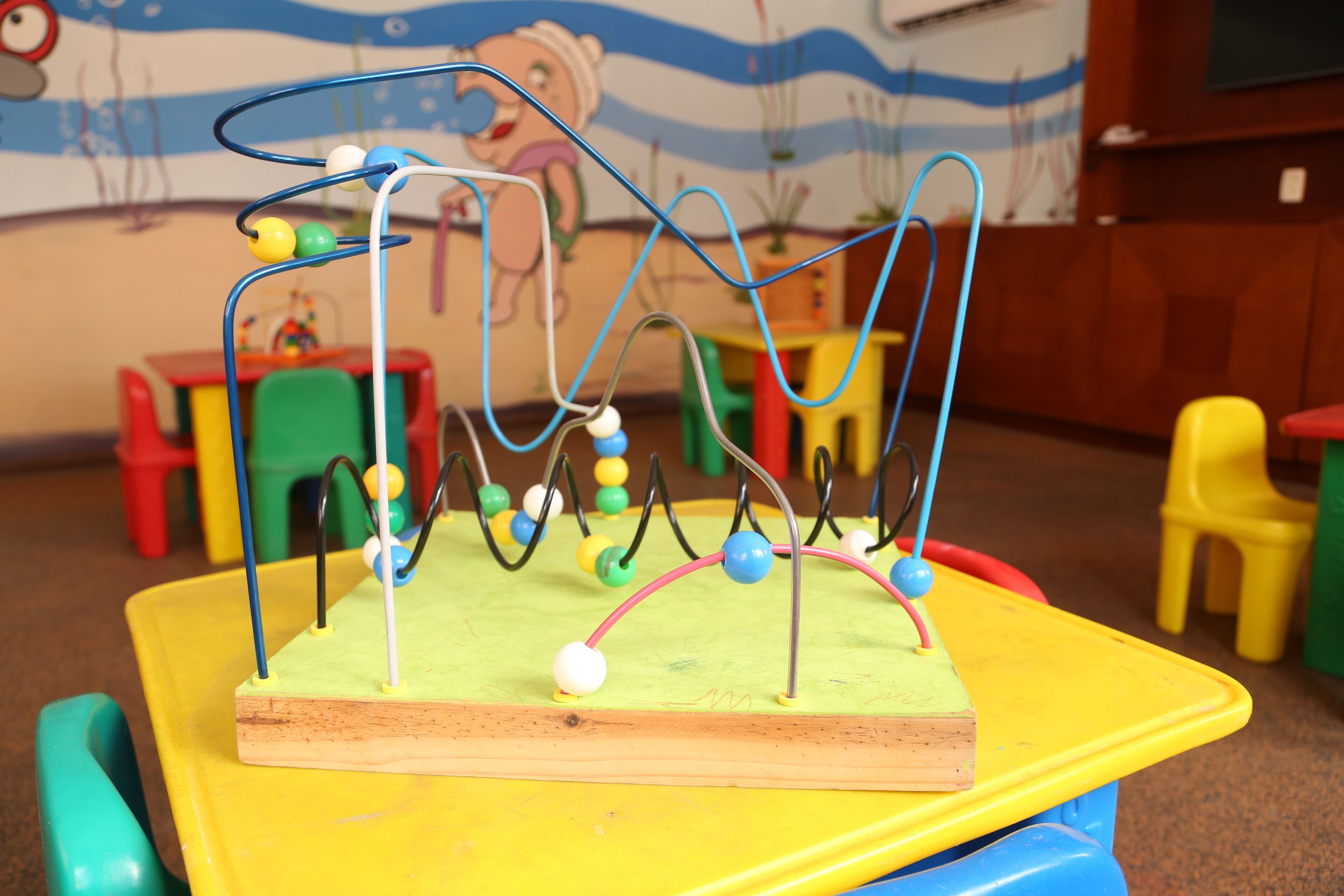 A colorful toy structure with various hanging elements on a wooden base, set against a wall with a cartoon character and a blue ocean-themed mural.