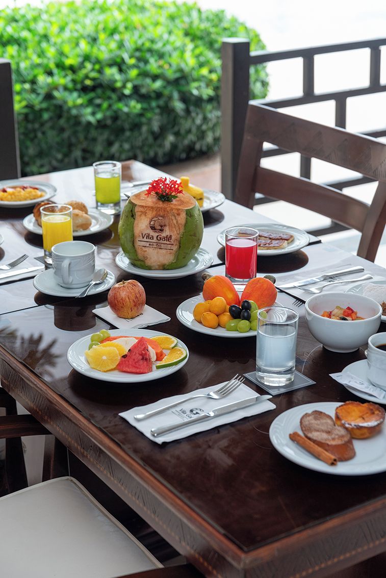 A breakfast table set with plates of fruit, a coconut, and beverages, with a view of greenery in the background.